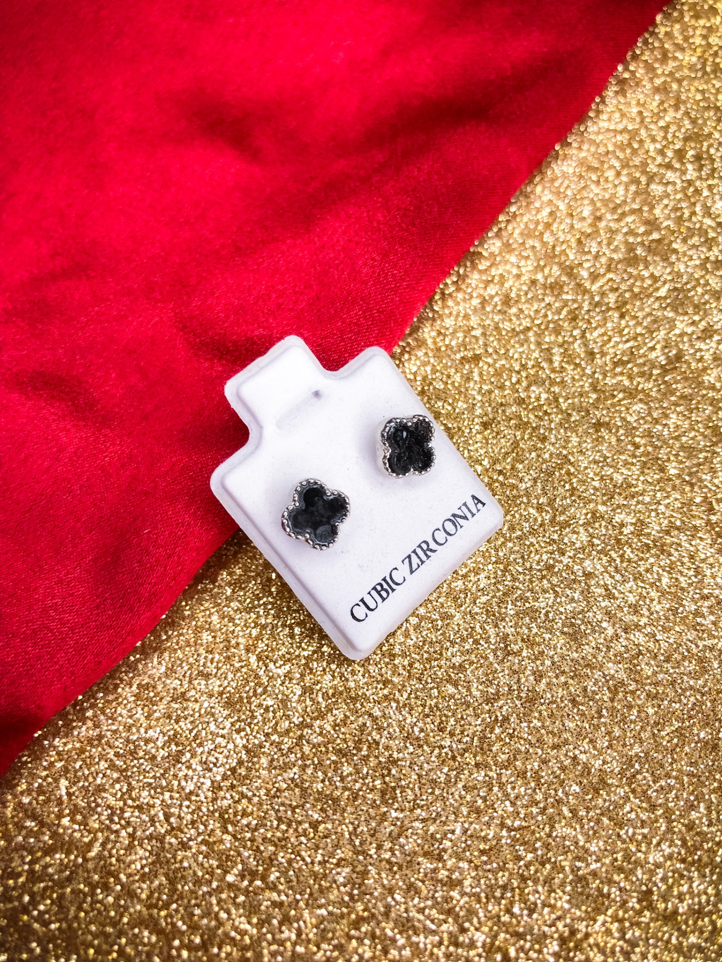 Brincos pretos em forma de flor sobre suporte branco com texto, em fundo dourado e vermelho