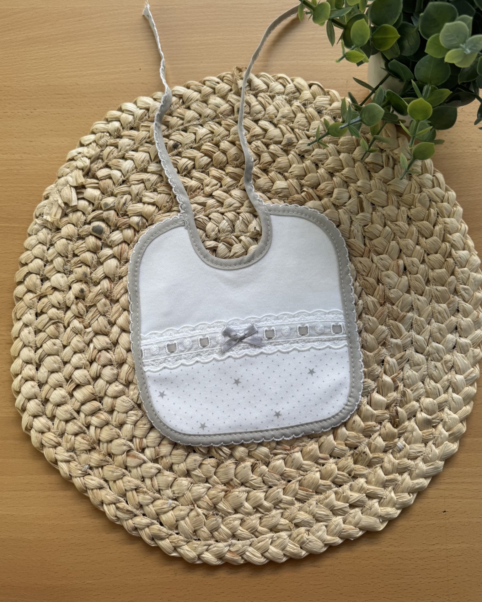 Babete de bebé branco e cinzento com padrão de estrelas pequenas e laço, sobre suporte trançado e planta verde.