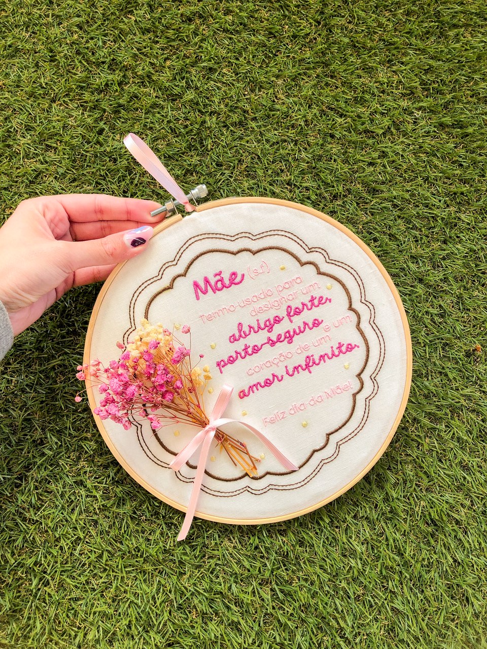 Aro de bordado com mensagem bordada e flores secas amarradas por fita rosa, mostrado sobre relva.