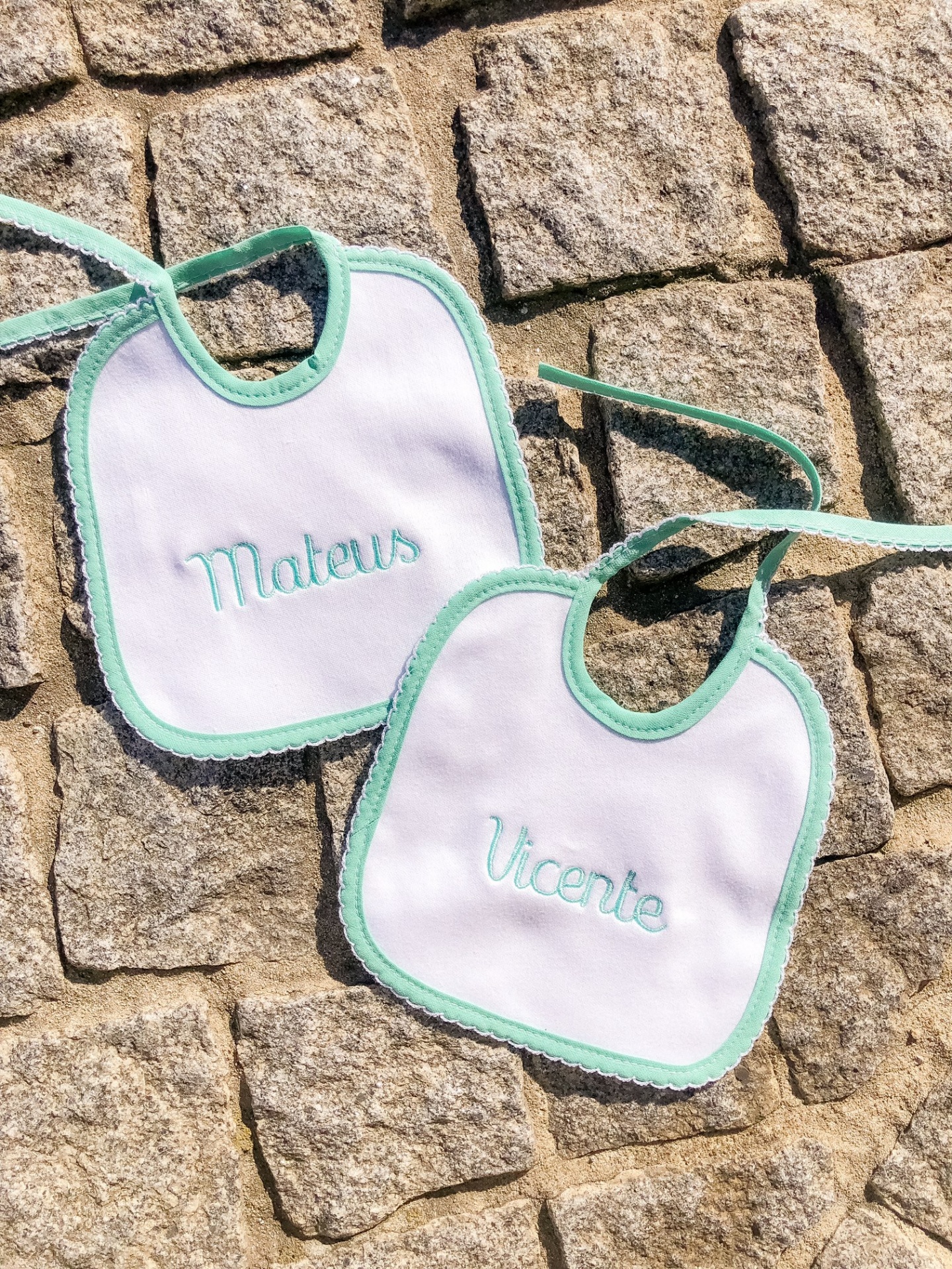 Dois babetes brancos com bordas verdes e nomes bordados colocados num chão de pedras