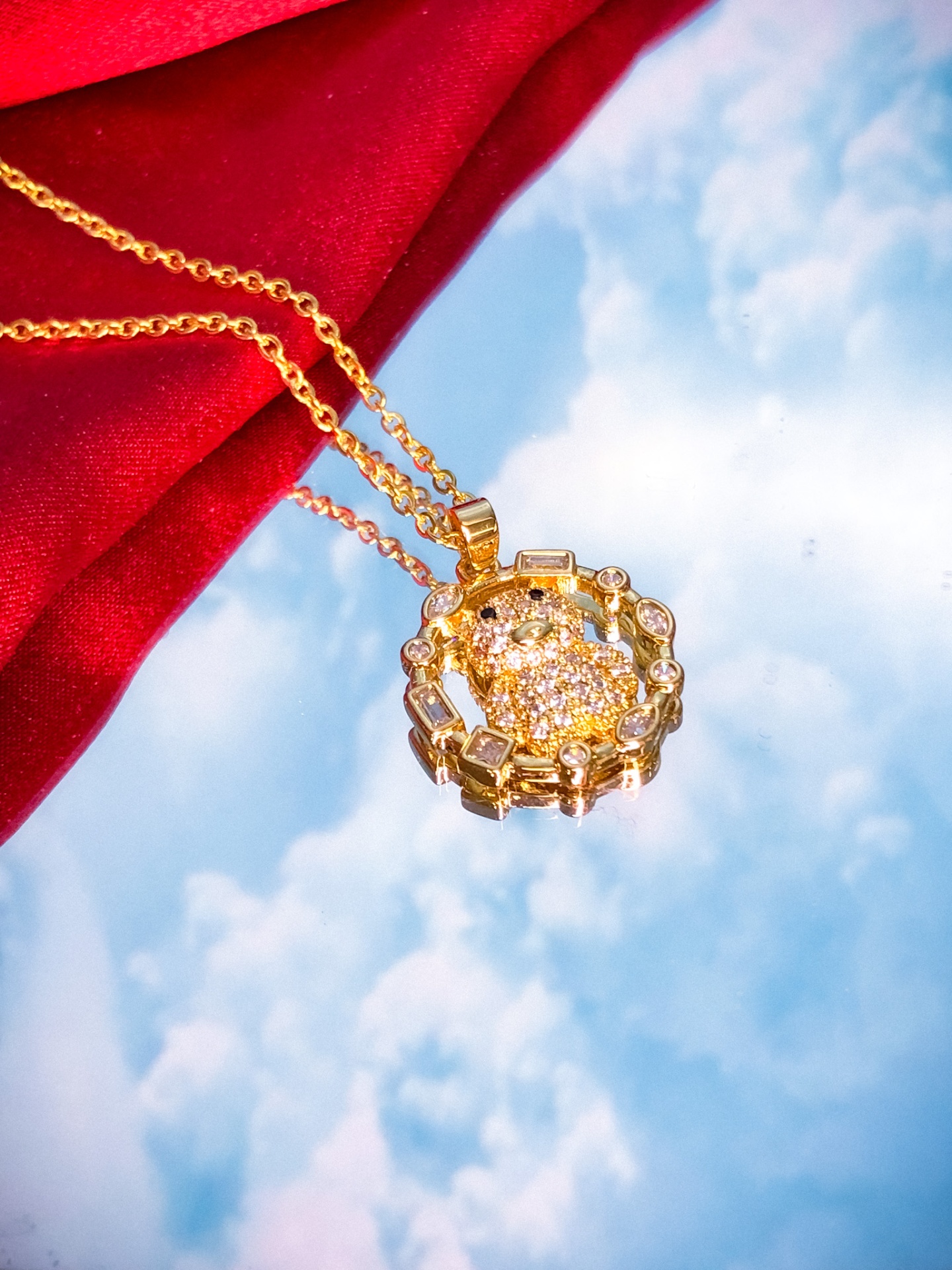 Colar dourado com pendente redondo com cristais sobre fundo de céu e tecido vermelho