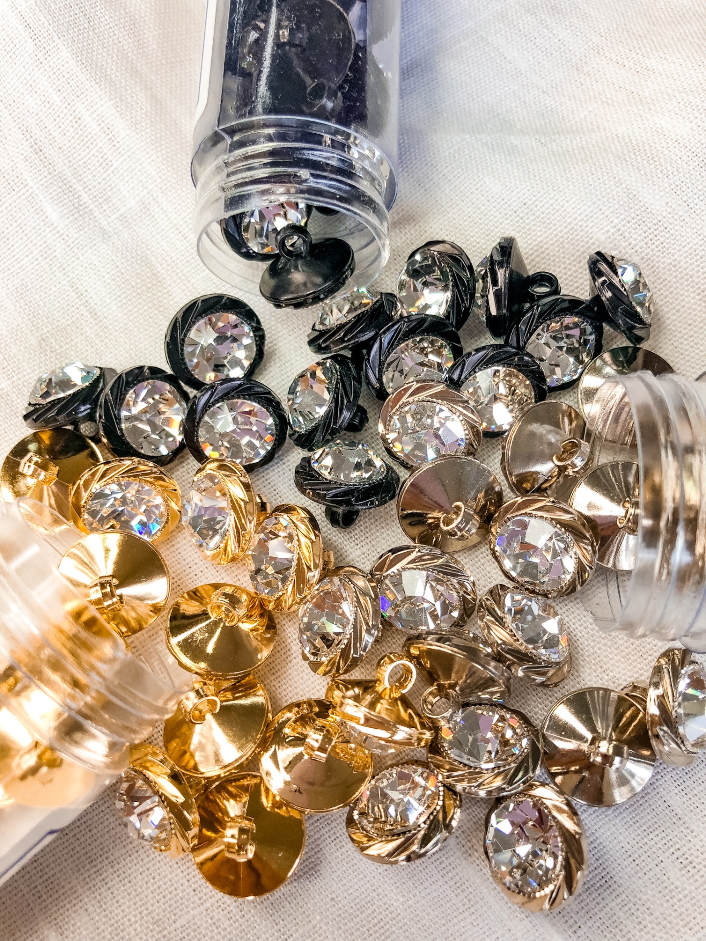 Botões decorativos dourados, prateados e pretos com cristais transparentes no centro, espalhados sobre tecido branco e frascos plásticos