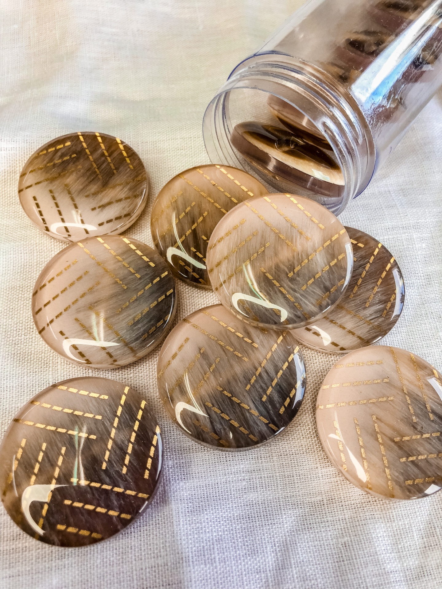 Botões redondos castanhos com padrão dourado sobre tecido branco e frasco plástico transparente