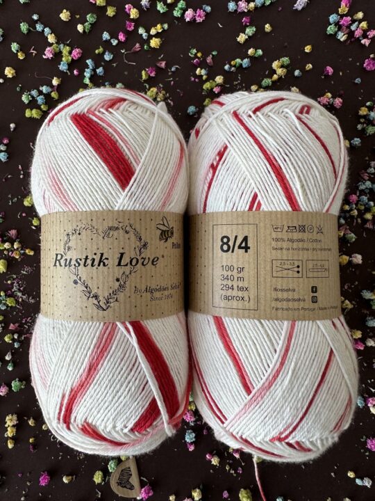 Dois novelos de fio de algodão branco com riscas vermelhas, rótulos castanhos, em fundo escuro com confetes coloridos