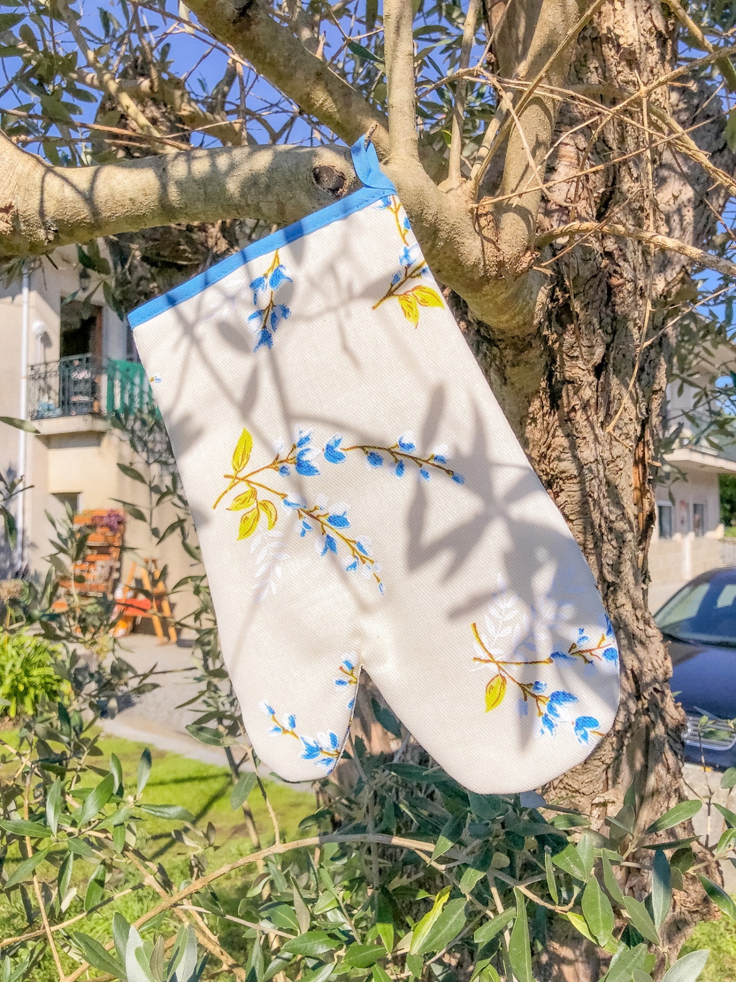 Luva de forno branca com padrão floral azul e amarelo pendurada numa árvore ao ar livre