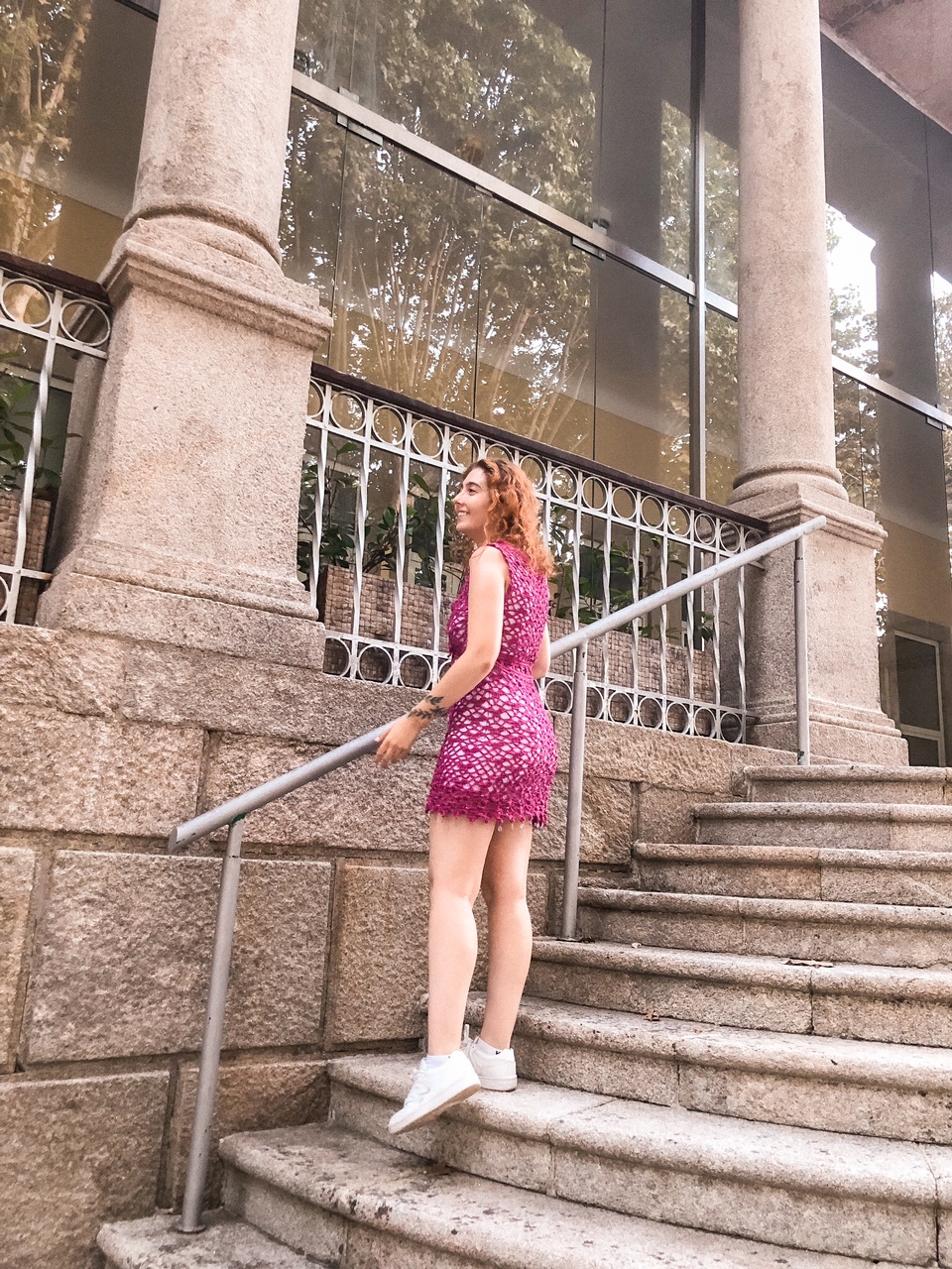 Mulher com vestido crochê rosa e ténis brancos numa escadaria de pedra com colunas.