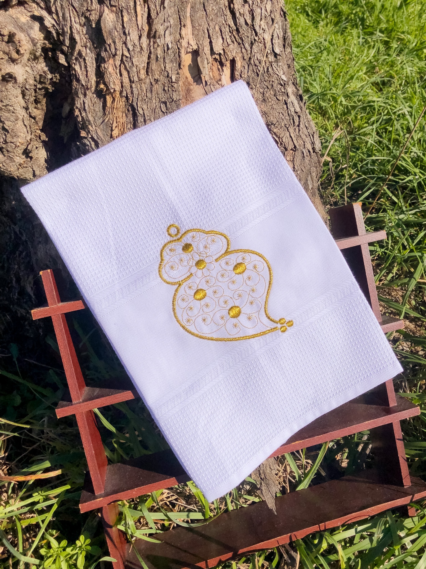 Toalha branca com bordado amarelo sobre estrutura de madeira junto a árvore e relva