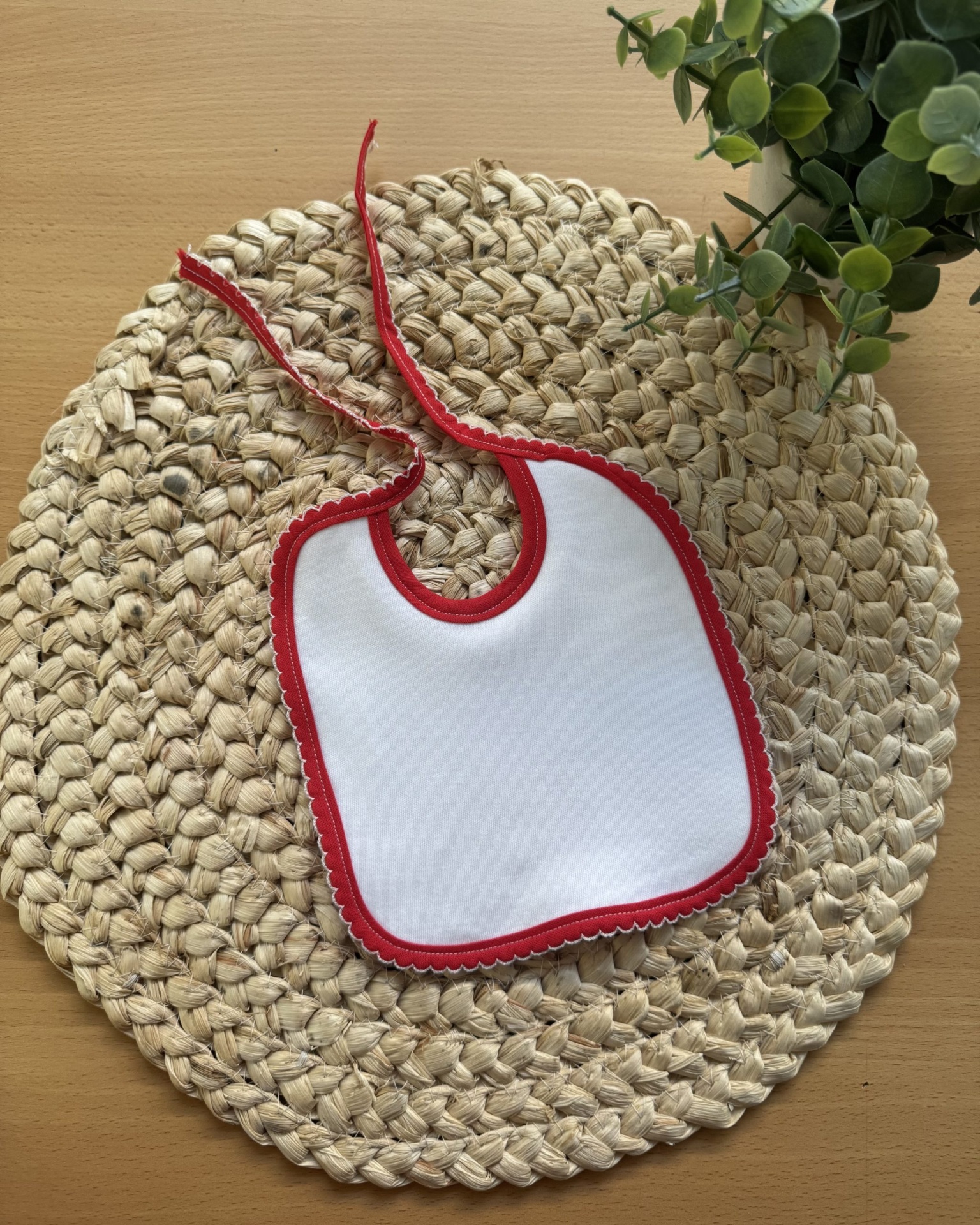 Babete branco com borda vermelha em tapete redondo de fibras naturais sobre mesa de madeira.