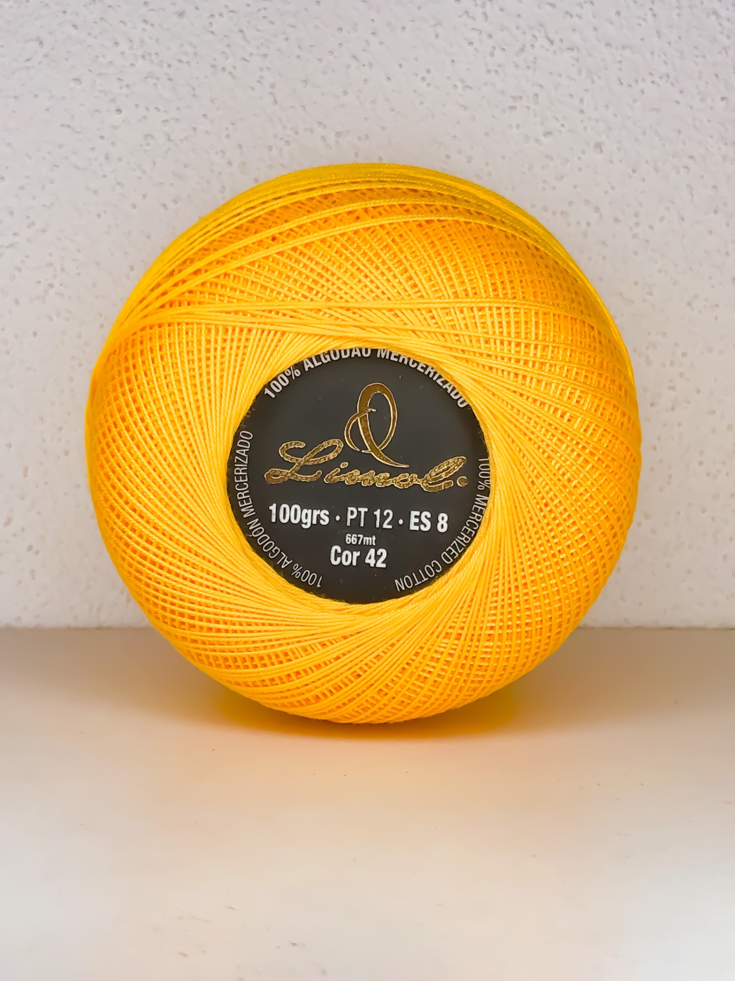 Novelo de linha amarela em algodão mercerizado com etiqueta preta e dourada, sobre superfície clara.