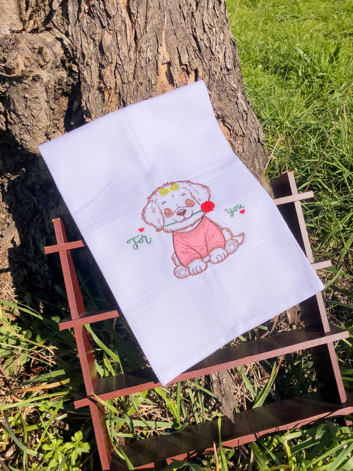 Toalha branca com bordado de cão sorridente e texto 'For You' encostada em árvore