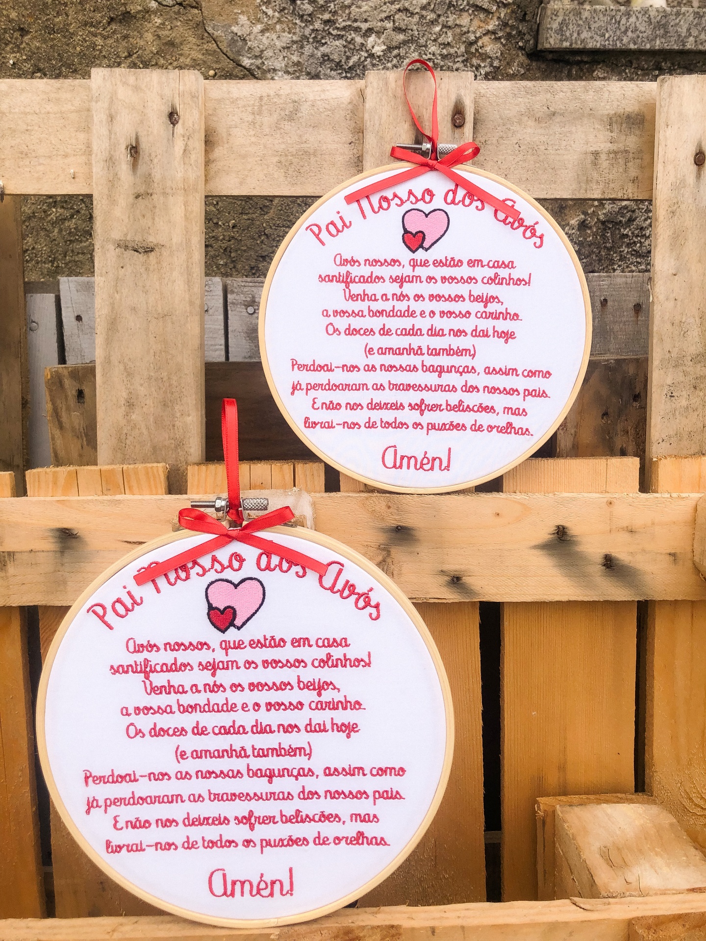 Dois painéis redondos em madeira com texto 'Pai Nosso dos Avós' em letras vermelhas, pendurados em fundo de madeira e pedra.