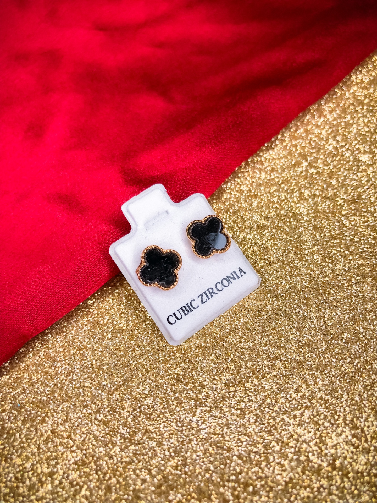 Brincos em forma de flor com pedras pretas em placa branca com texto, sobre superfície dourada e pano vermelho