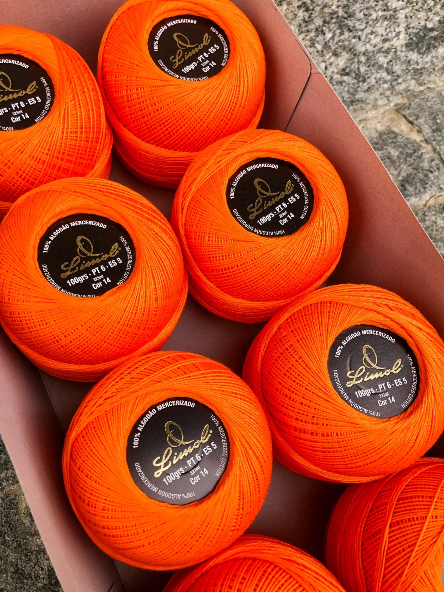 Oito novelos de linha laranja 100% algodão mercerizado com rótulo preto e dourado dentro de uma caixa castanha