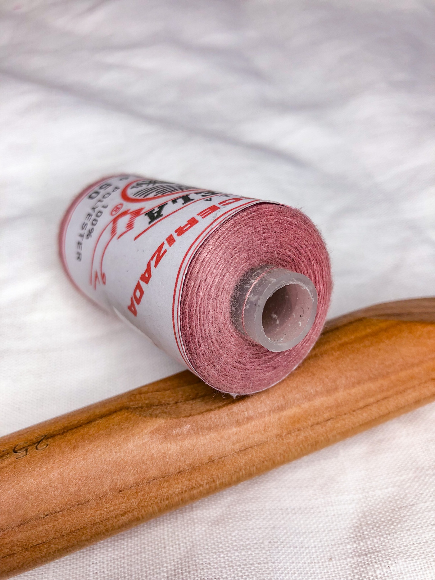 Rolo de linha cor-de-rosa com rótulo e agulha de madeira sobre tecido branco.