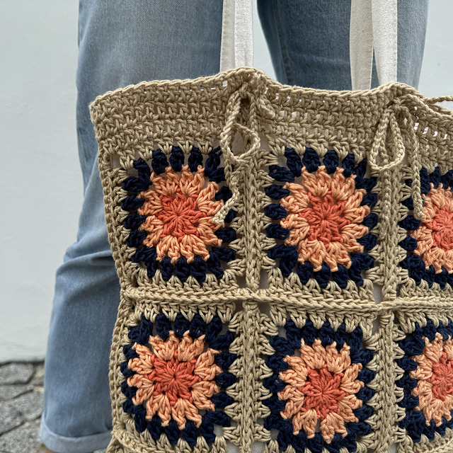 Mala tricotada em quadrados coloridos, com alças de tecido, segurada por pessoa com calças azuis