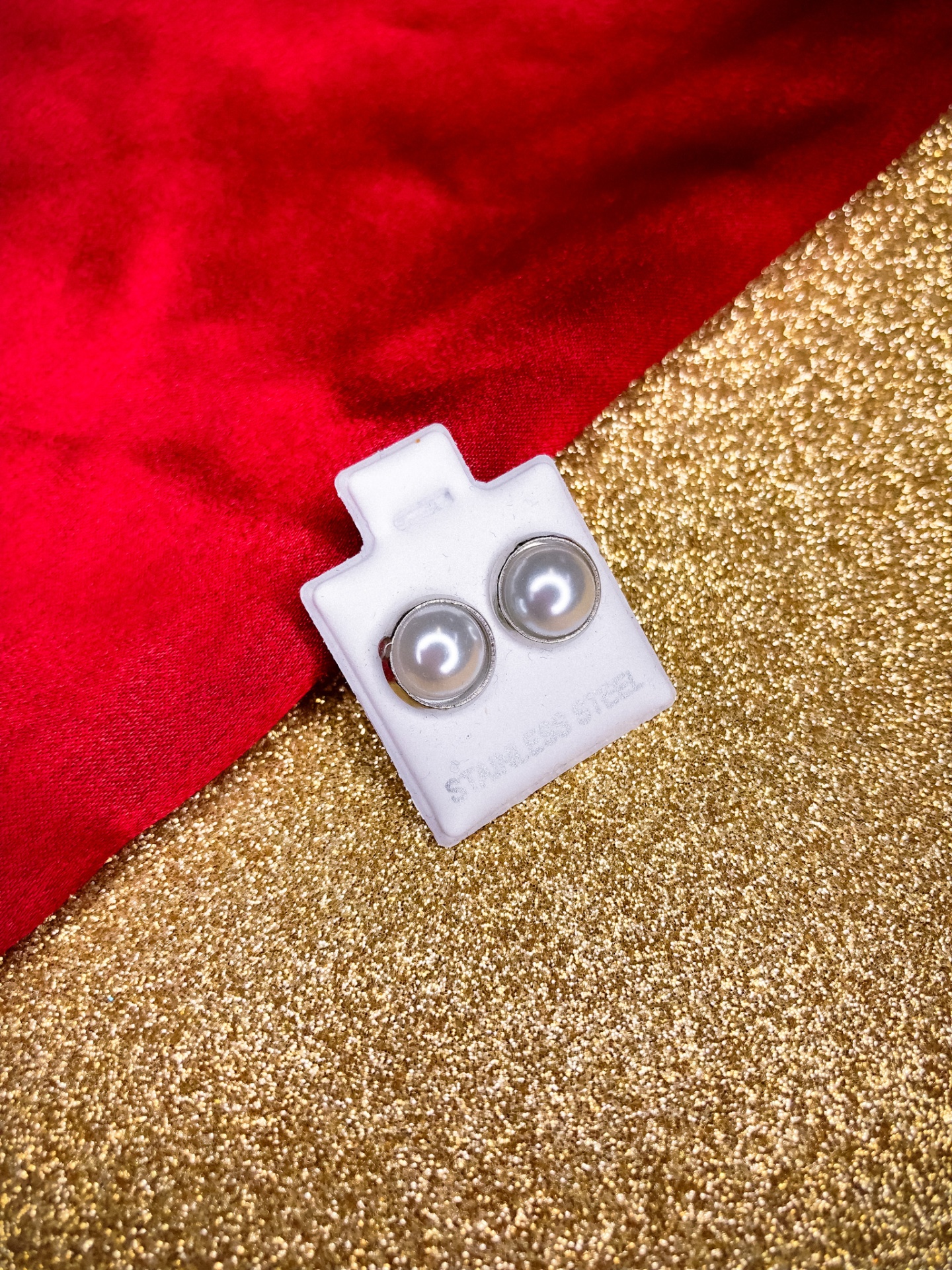 Brincos de pérola cinzenta em aço inoxidável num suporte branco sobre fundo vermelho e dourado