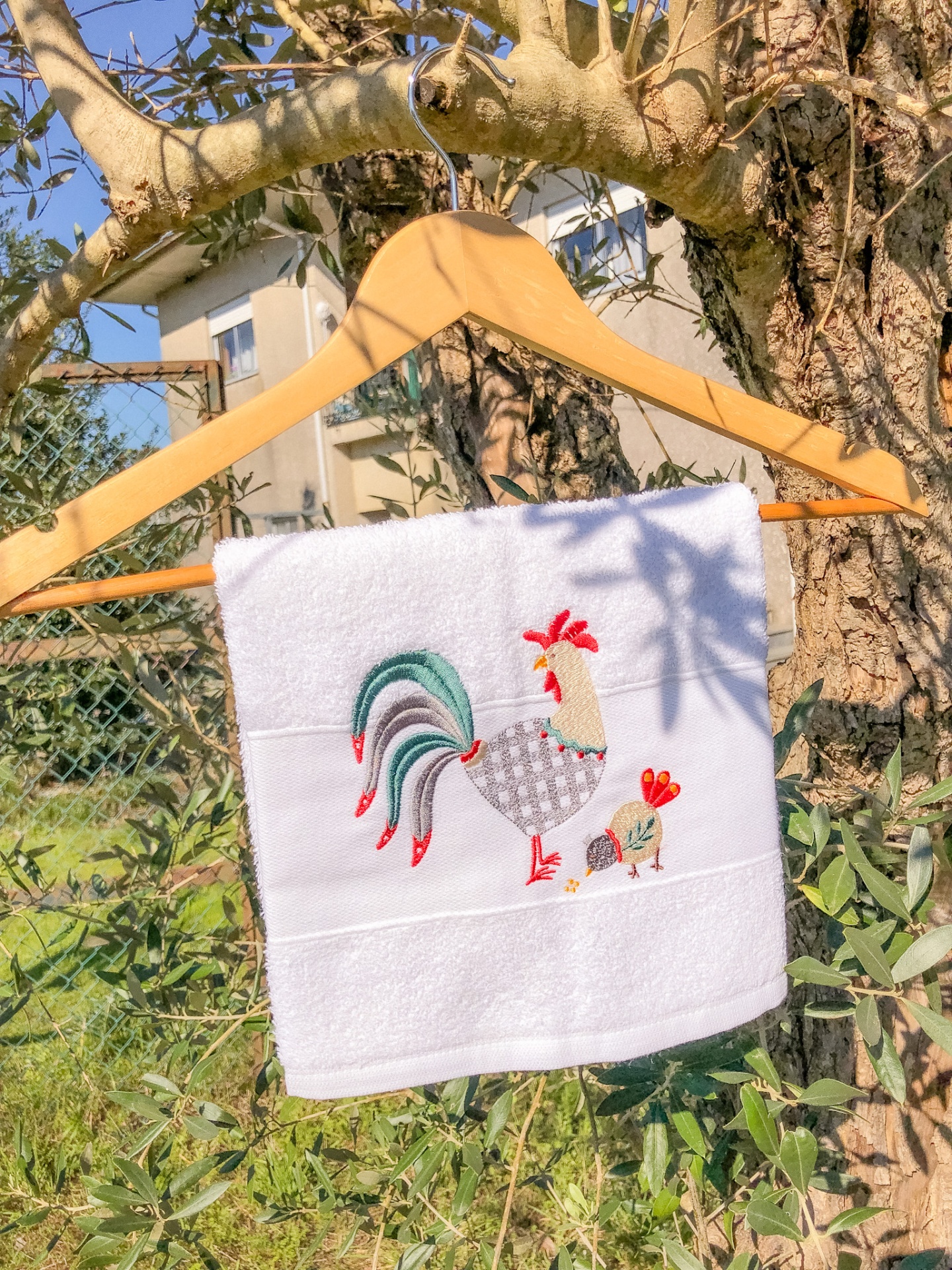 Toalha branca com bordado de galo e pintainho pendurada num cabide de madeira numa árvore