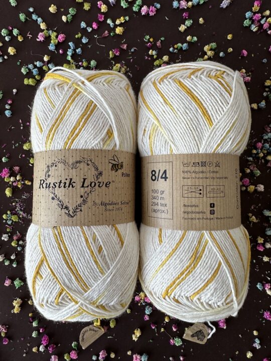 Dois novelos de lã branca e amarela Rustik Love com etiquetas de papel kraft sobre fundo escuro com flores secas coloridas.