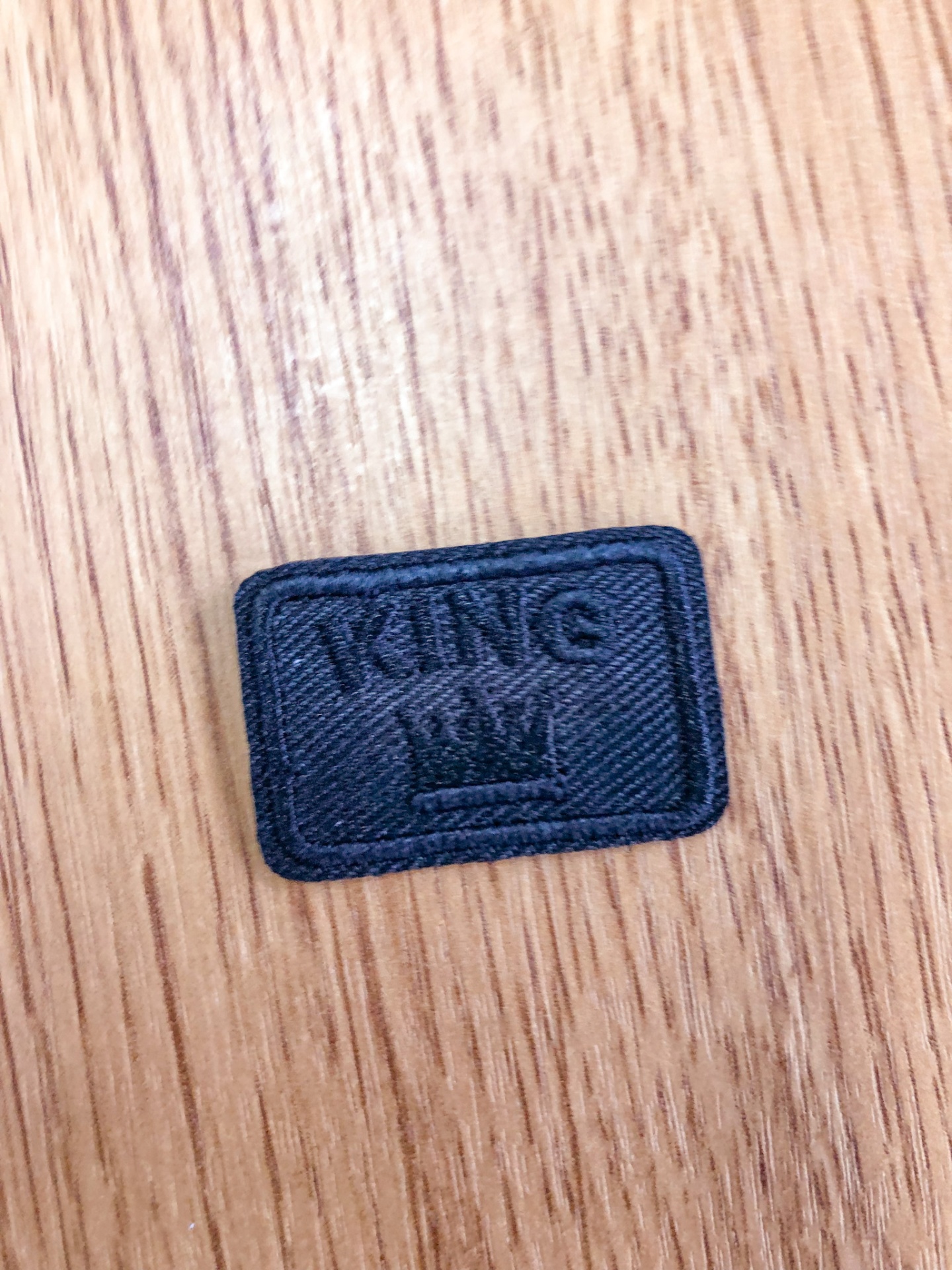 Etiqueta preta de tecido com a palavra KING e coroa sobre superfície de madeira.