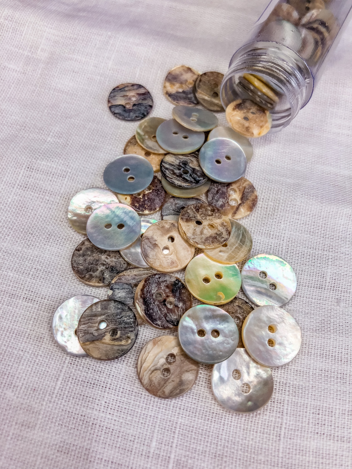 Botões redondos em tons naturais e iridescentes com dois furos, sobre pano branco, junto a um frasco de vidro