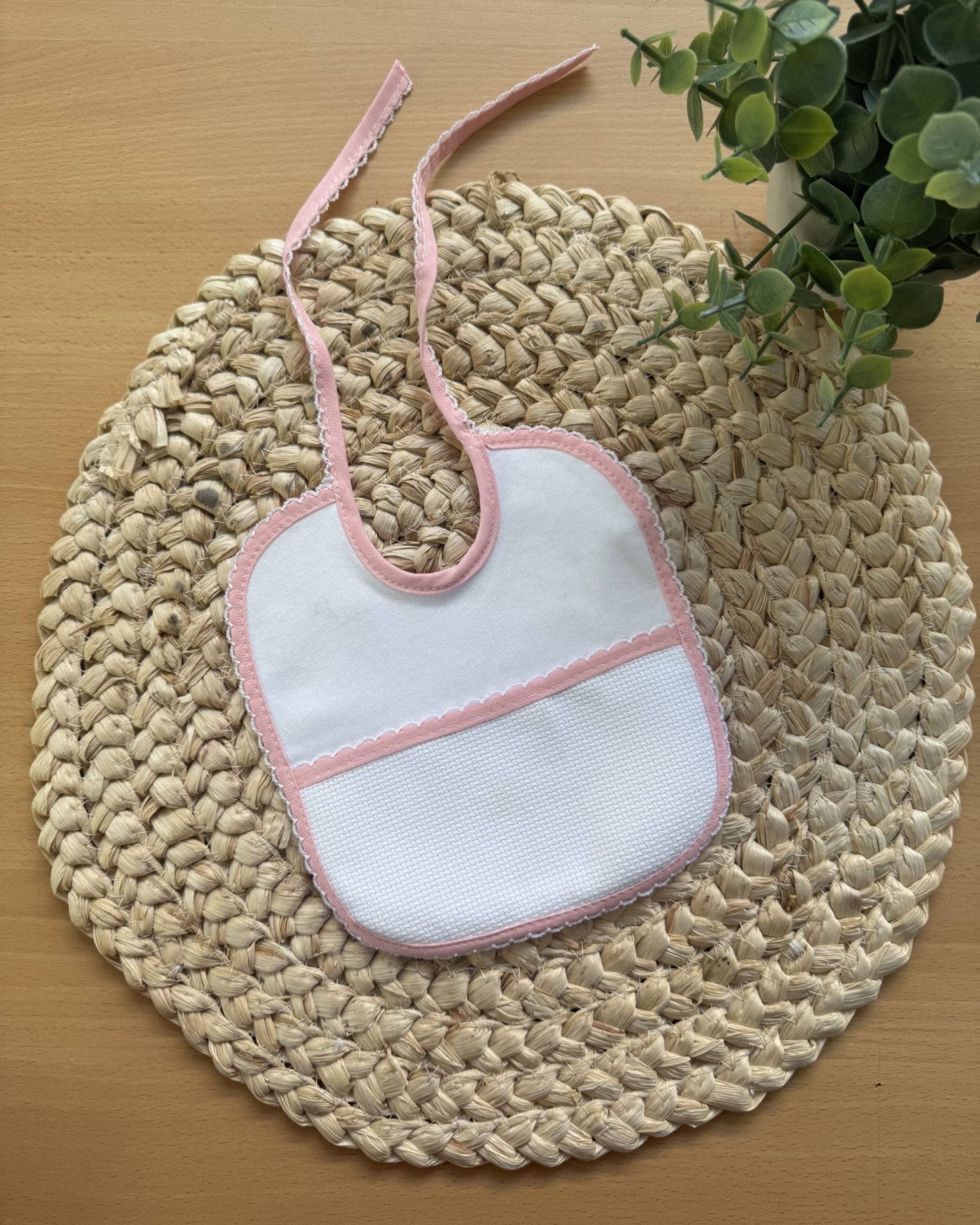 Babete branco com bordas cor-de-rosa em tapete de palha redondo, com planta verde ao lado.