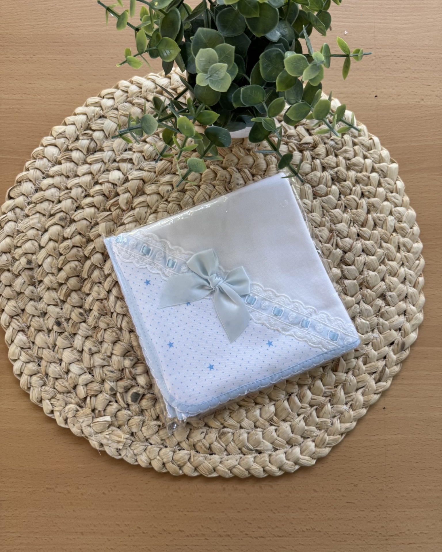 Toalha branca com bordado azul forte, renda e fita, sobre tapete redondo trançado e planta artificial