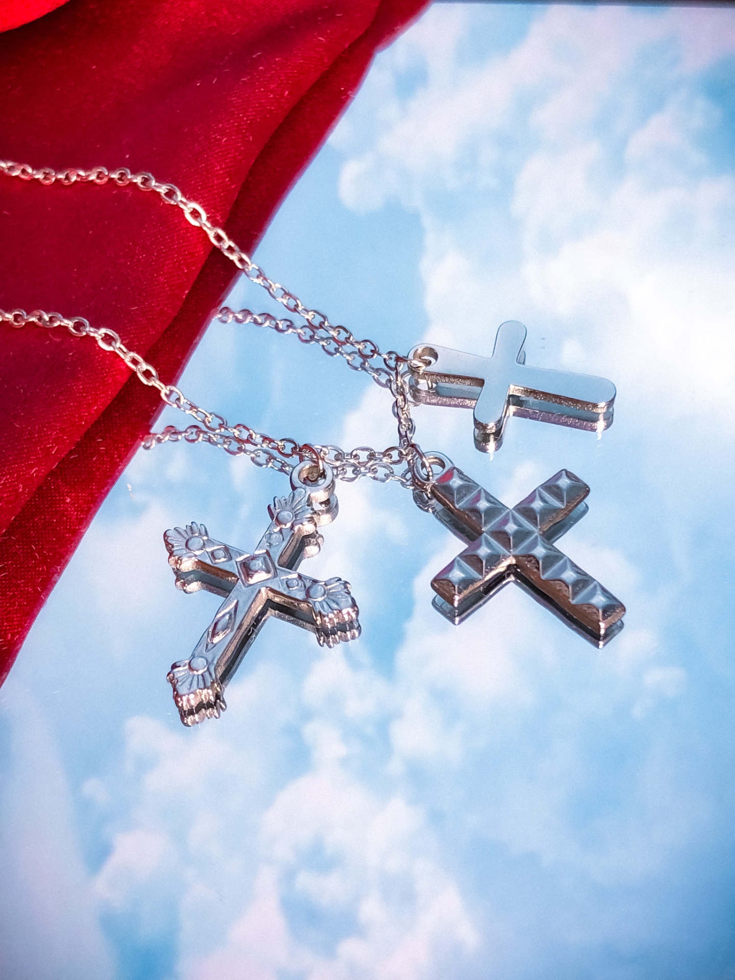 Colares com três cruzes em metal prateado sobre fundo azul e tecido vermelho