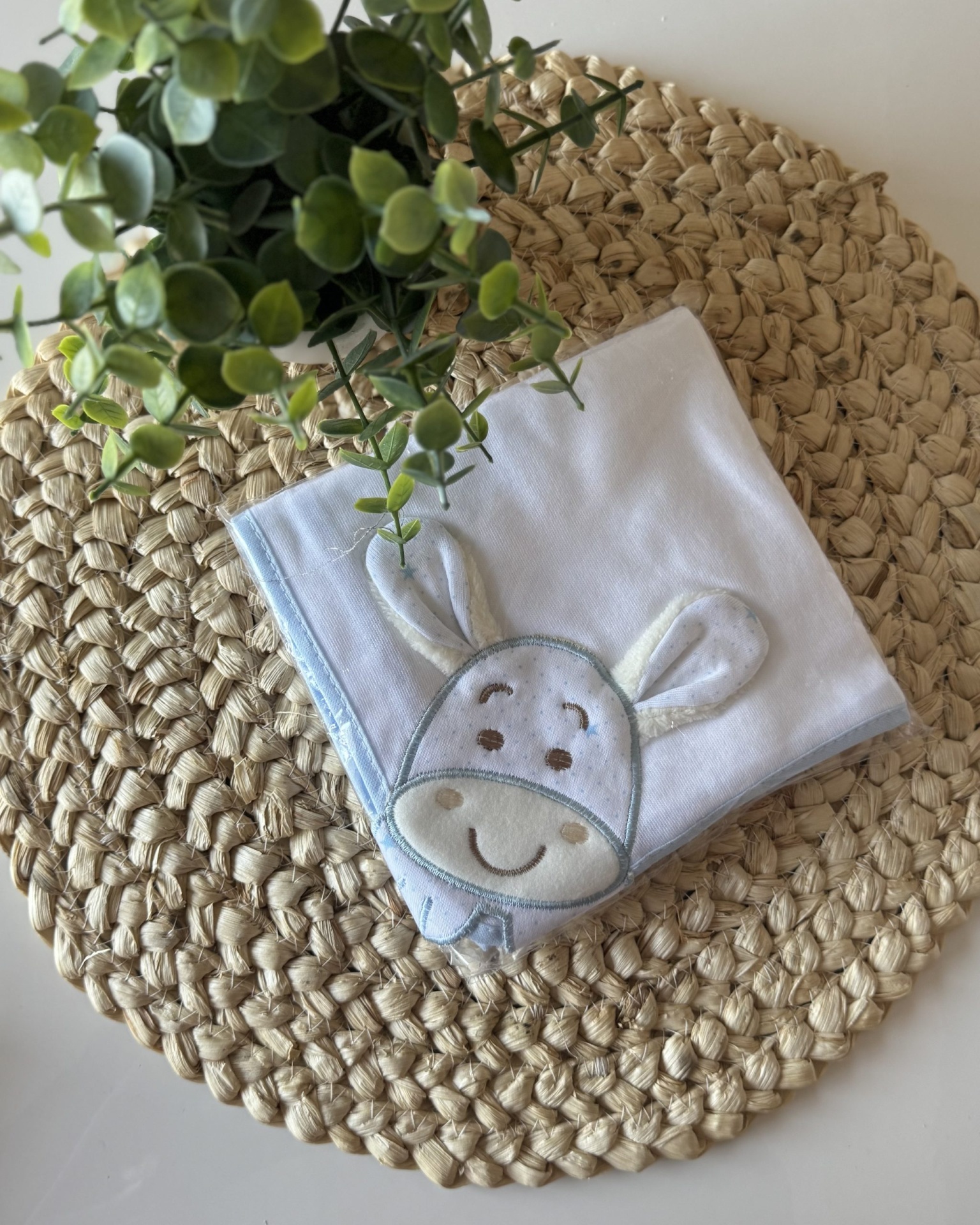 Fralda branca com bordado de rosto de burro sobre suporte trançado com folhas verdes
