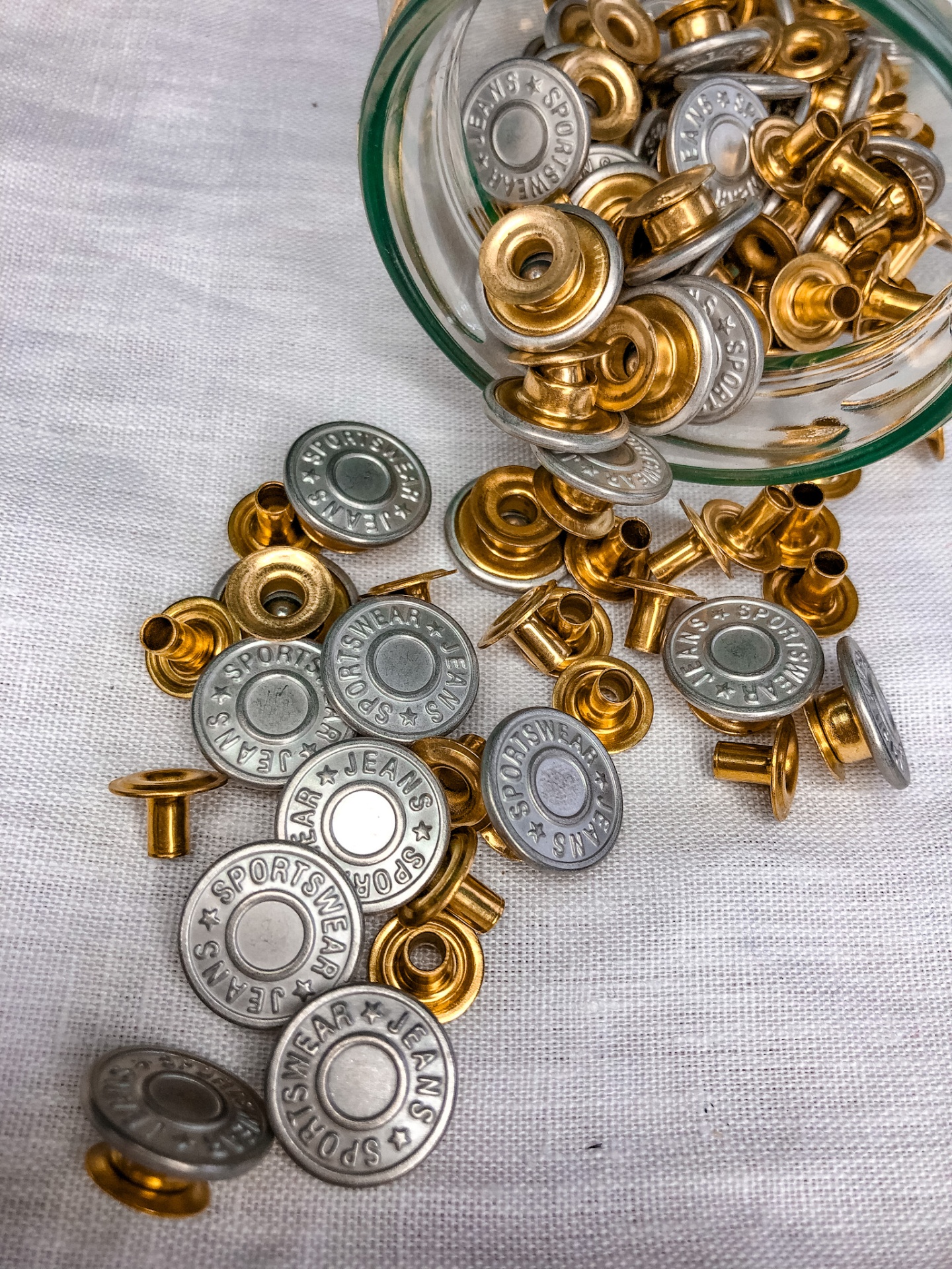 Botões metálicos prateados e dourados com gravação, sobre pano branco