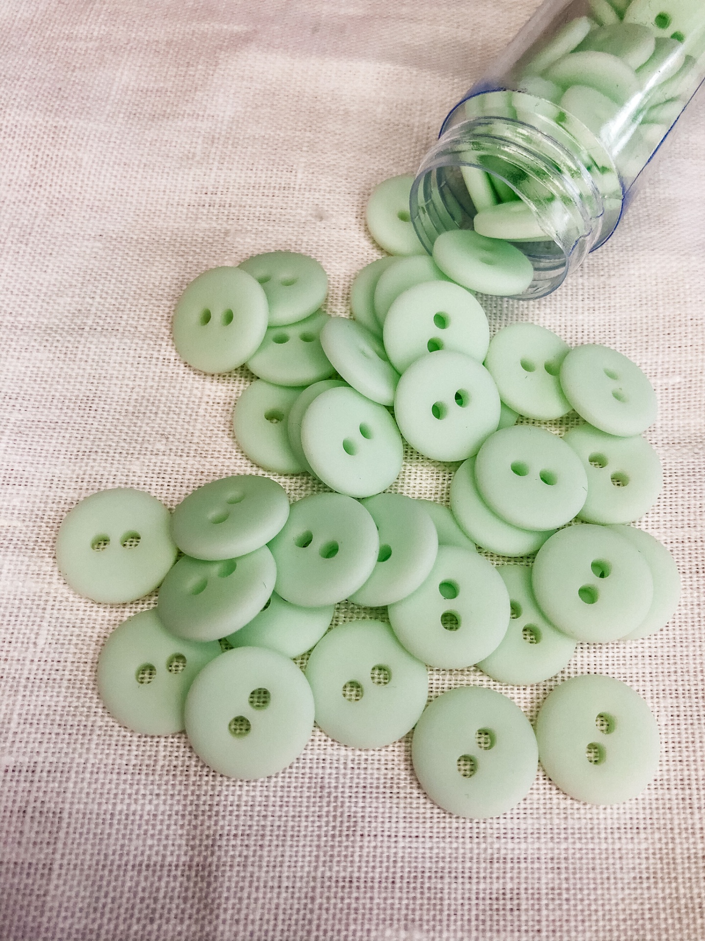 Botões verdes claros com dois furos, sobre tecido bege, e tubo plástico transparente ao fundo.