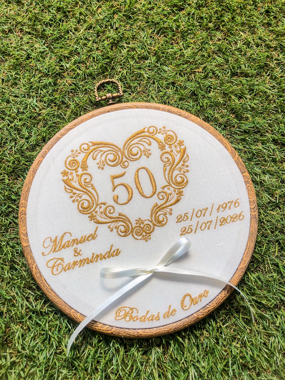 Bastidor bordado redondo branco com texto em dourado e fita branca sobre relva