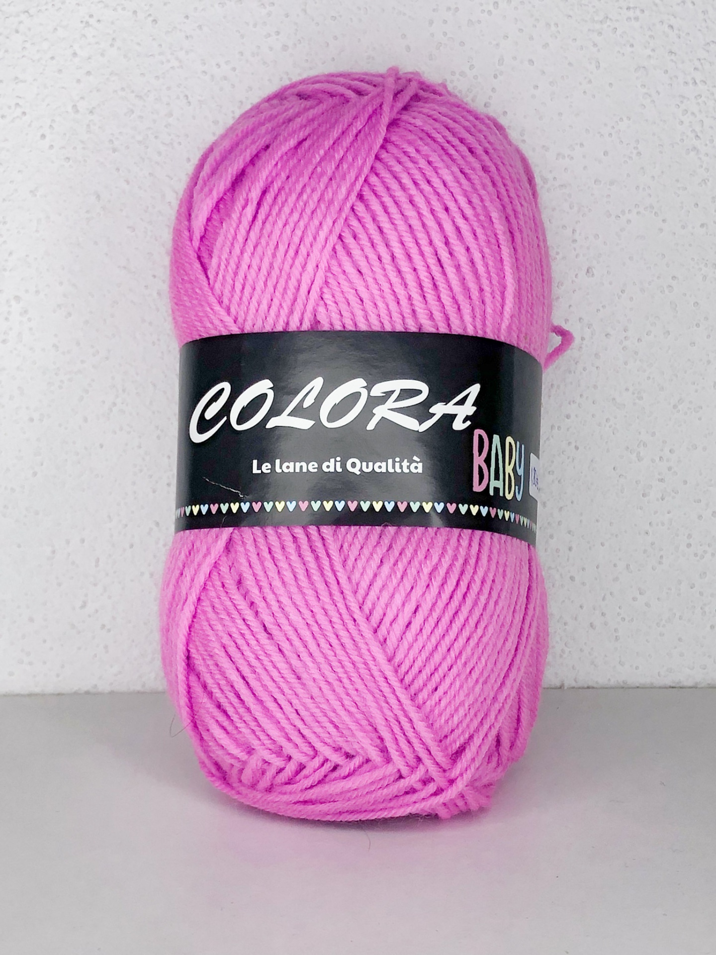 Novelo de lã cor rosa com etiqueta preta e texto branco e colorido, sobre superfície cinza claro com fundo branco