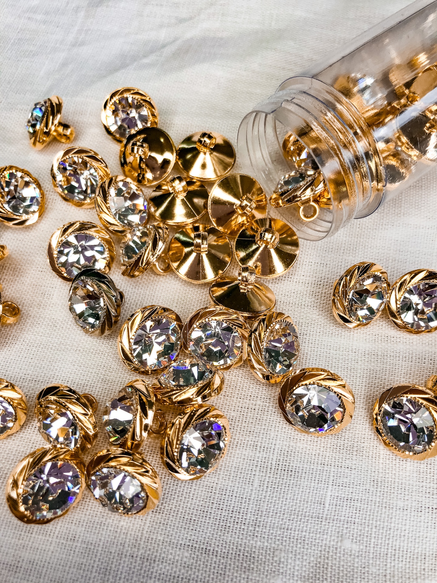 Botões dourados com pedras facetadas sobre tecido branco e frasco plástico