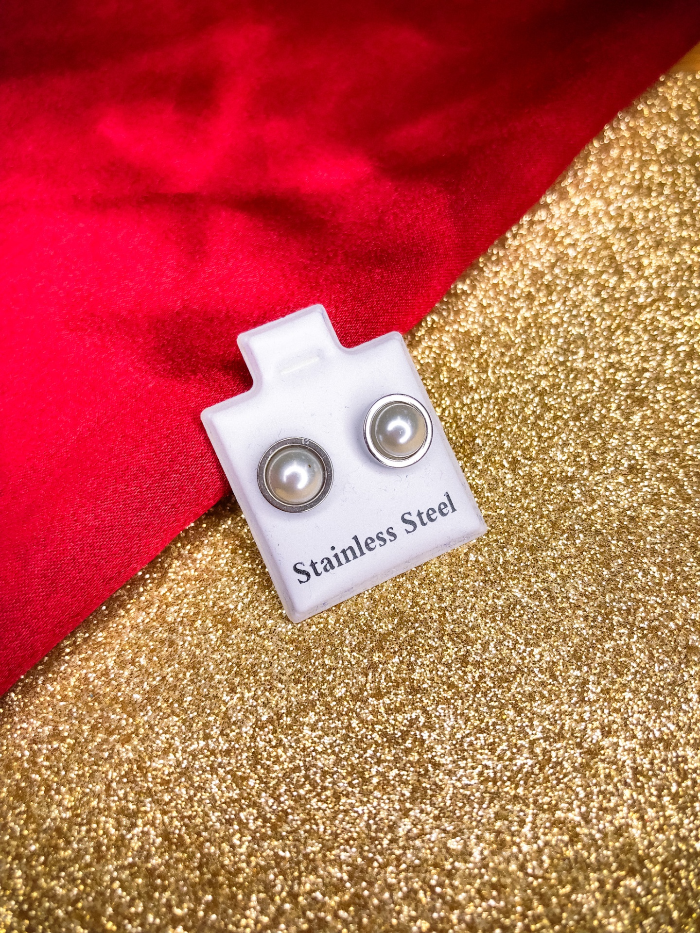 Brincos pérola em aço inoxidável em suporte branco com texto sobre fundo dourado e tecido vermelho