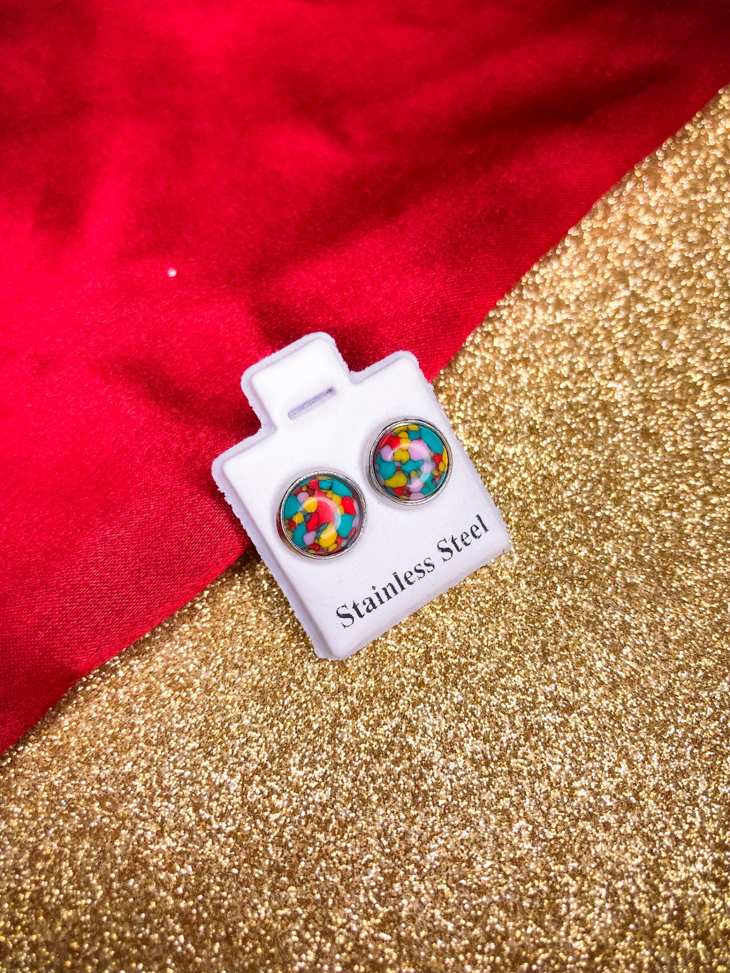 Brincos redondos coloridos em aço inoxidável sobre suporte branco com texto, fundo vermelho e dourado.