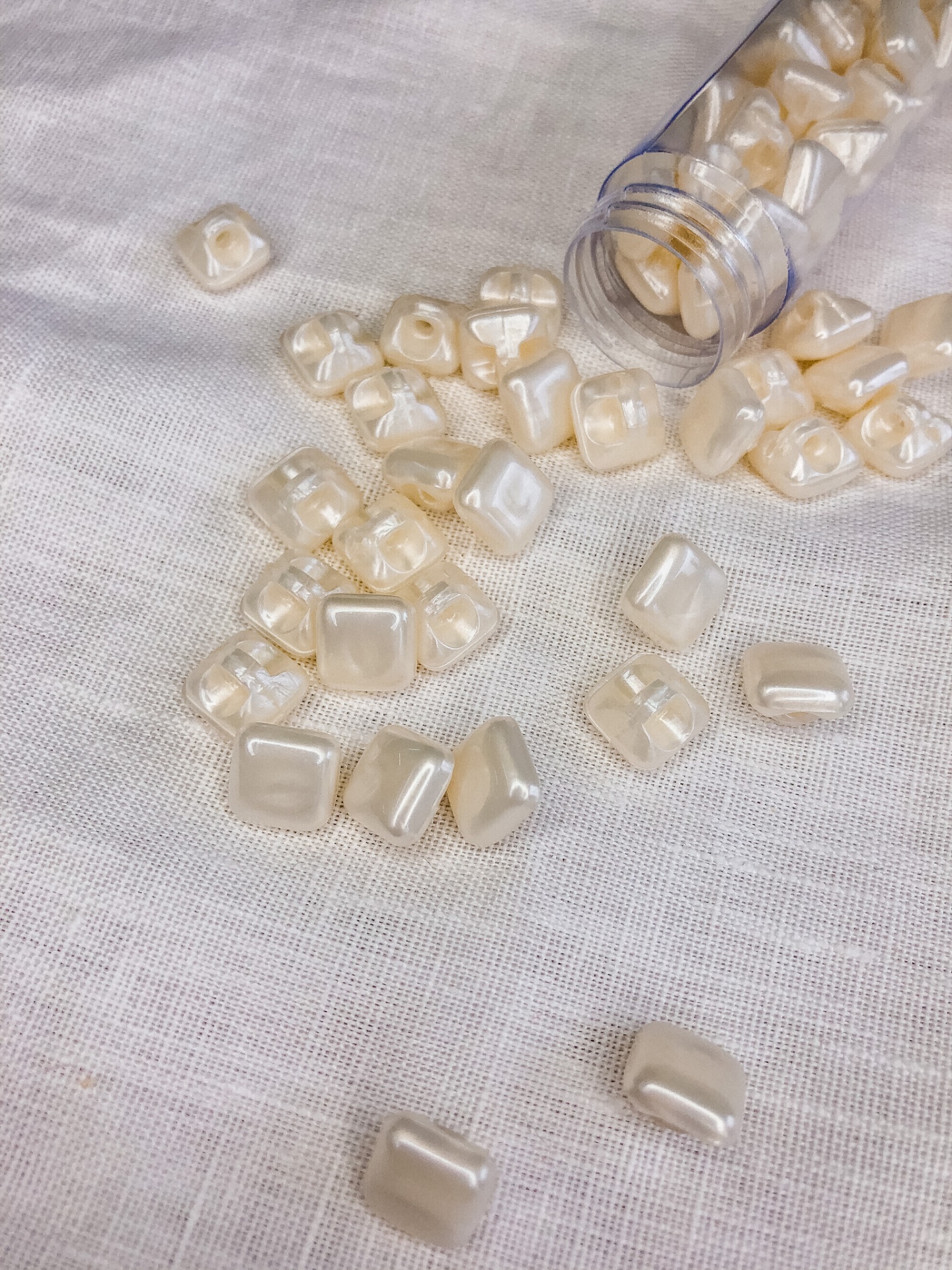 Contas quadradas pérola plásticas a sair de frasco sobre tecido branco