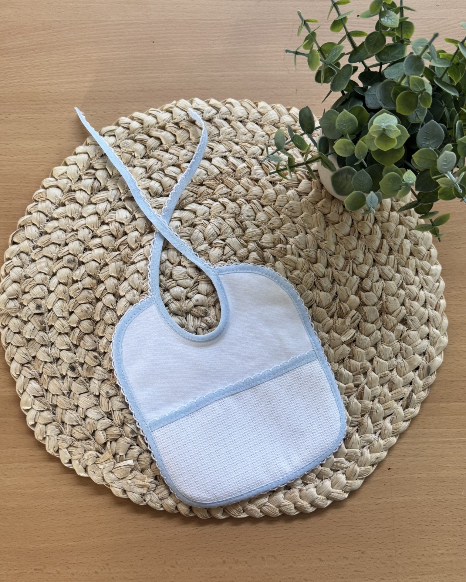 Babete azul e branco para bebé sobre base de ráfia com planta verde ao lado
