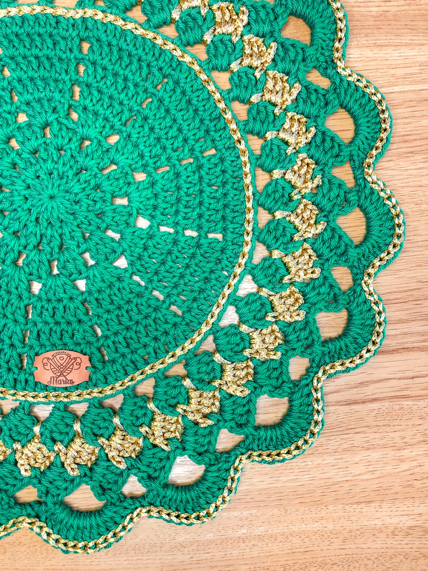 Toalha redonda verde e dourada de crochê com etiqueta MARIA em fundo de madeira
