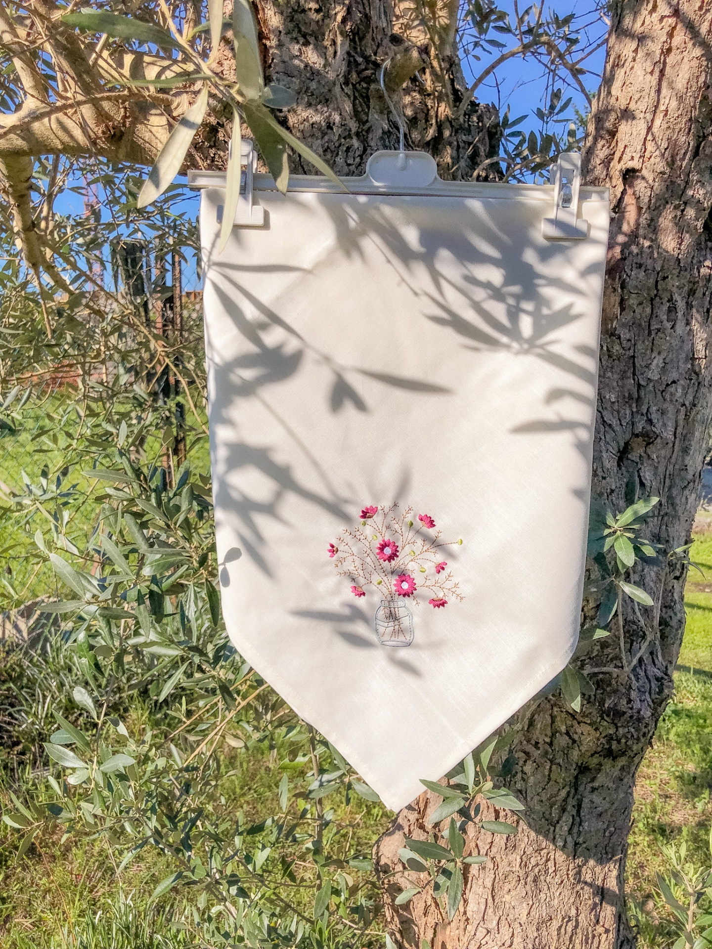 Pano branco com desenho de vaso de flores pendurado numa árvore de oliveira