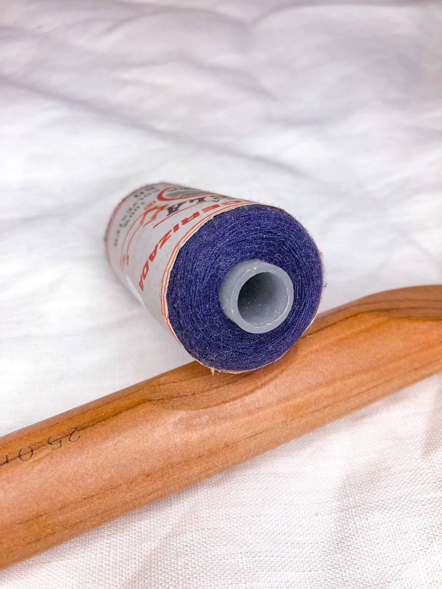 Rolo de linha roxa com rótulo em tecido branco junto de alfinete de madeira.