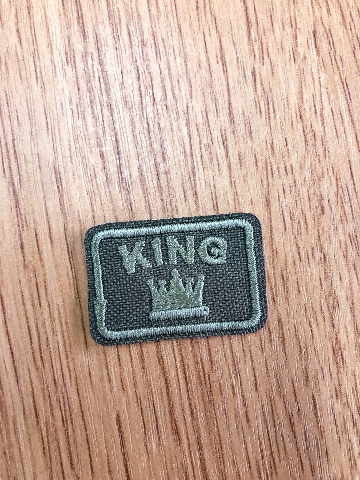 Emblema bordado cinza escuro com texto KING e coroa sobre madeira clara