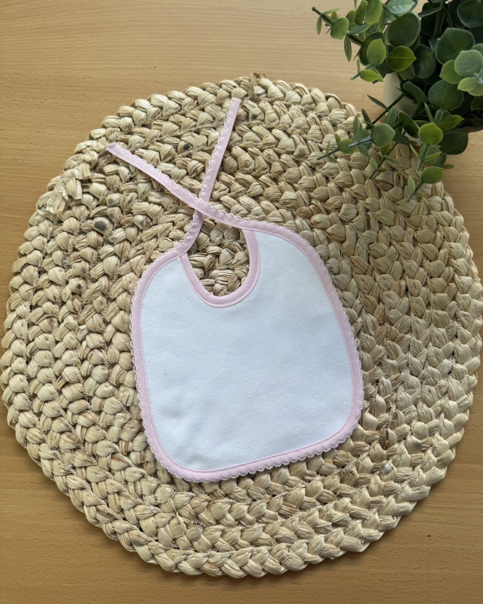 Babete branco com borda e cordões cor-de-rosa em base redonda de corda trançada clara, com plantas verdes.