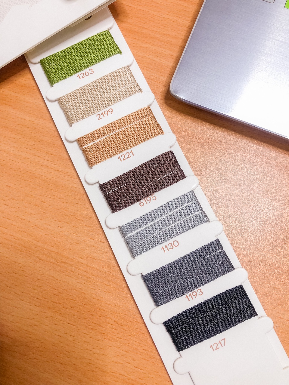 Mostruário de tecidos coloridos numerados em diferentes tons sobre mesa de madeira ao lado de portátil prateado