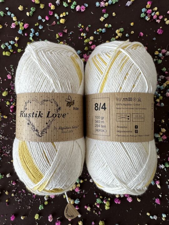 Dois novelos de lã Rustik Love branco e amarelo com etiquetas castanhas, sobre fundo escuro com flores secas coloridas.