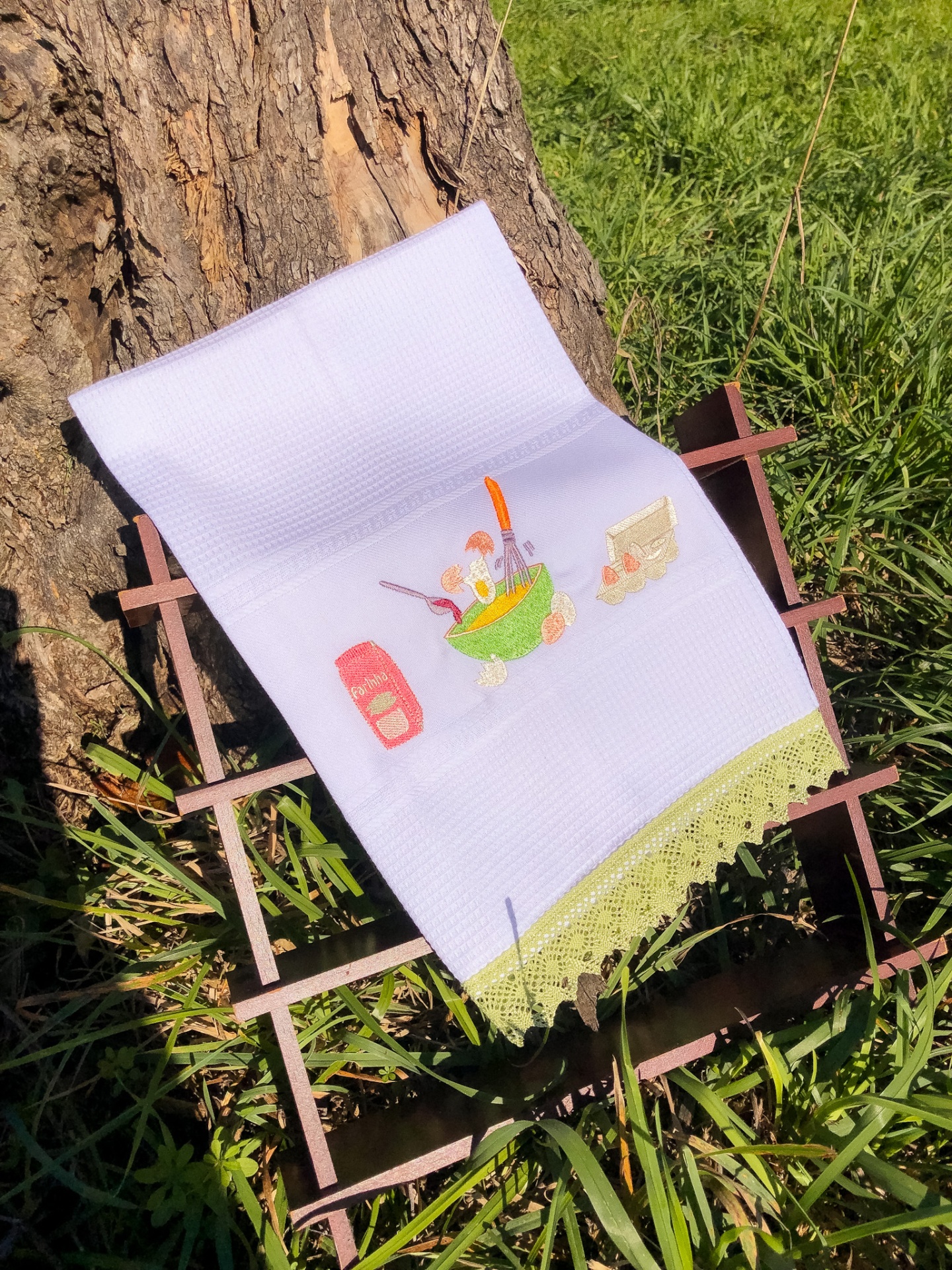 Pano de cozinha branco com bordado de tigela e renda verde clara sobre suporte de madeira ao ar livre