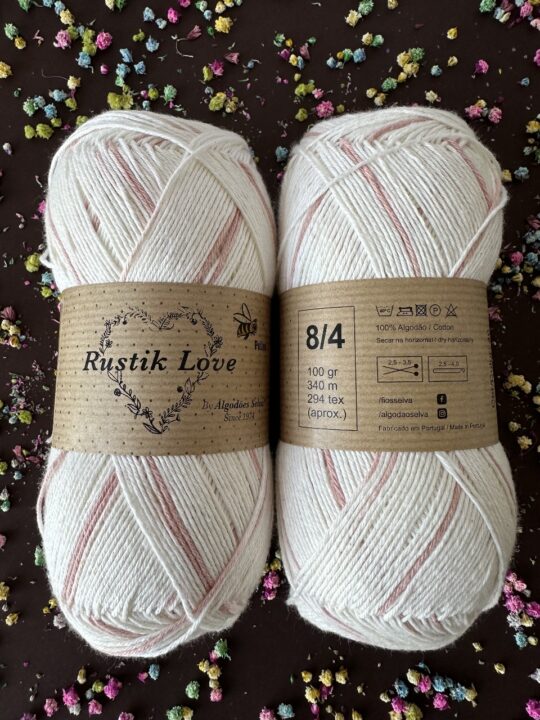 Duas meadas de lã branca com riscas rosas e rótulo castanho Rustik Love sobre fundo escuro com pequenos corpos coloridos.