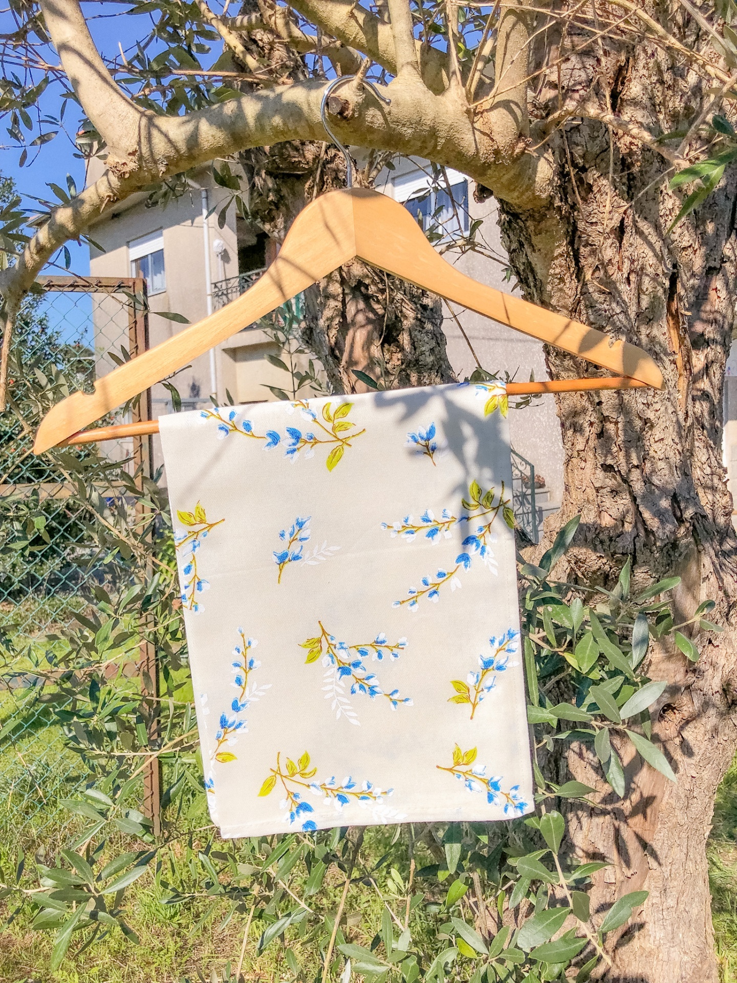 Lenço branco com flores azuis e folhas verdes pendurado num cabide de madeira numa árvore