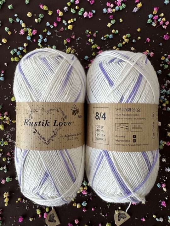 Duas meadas de lã Rustik Love branco com detalhes lilás e rótulos castanhos, sobre fundo com flores coloridas.