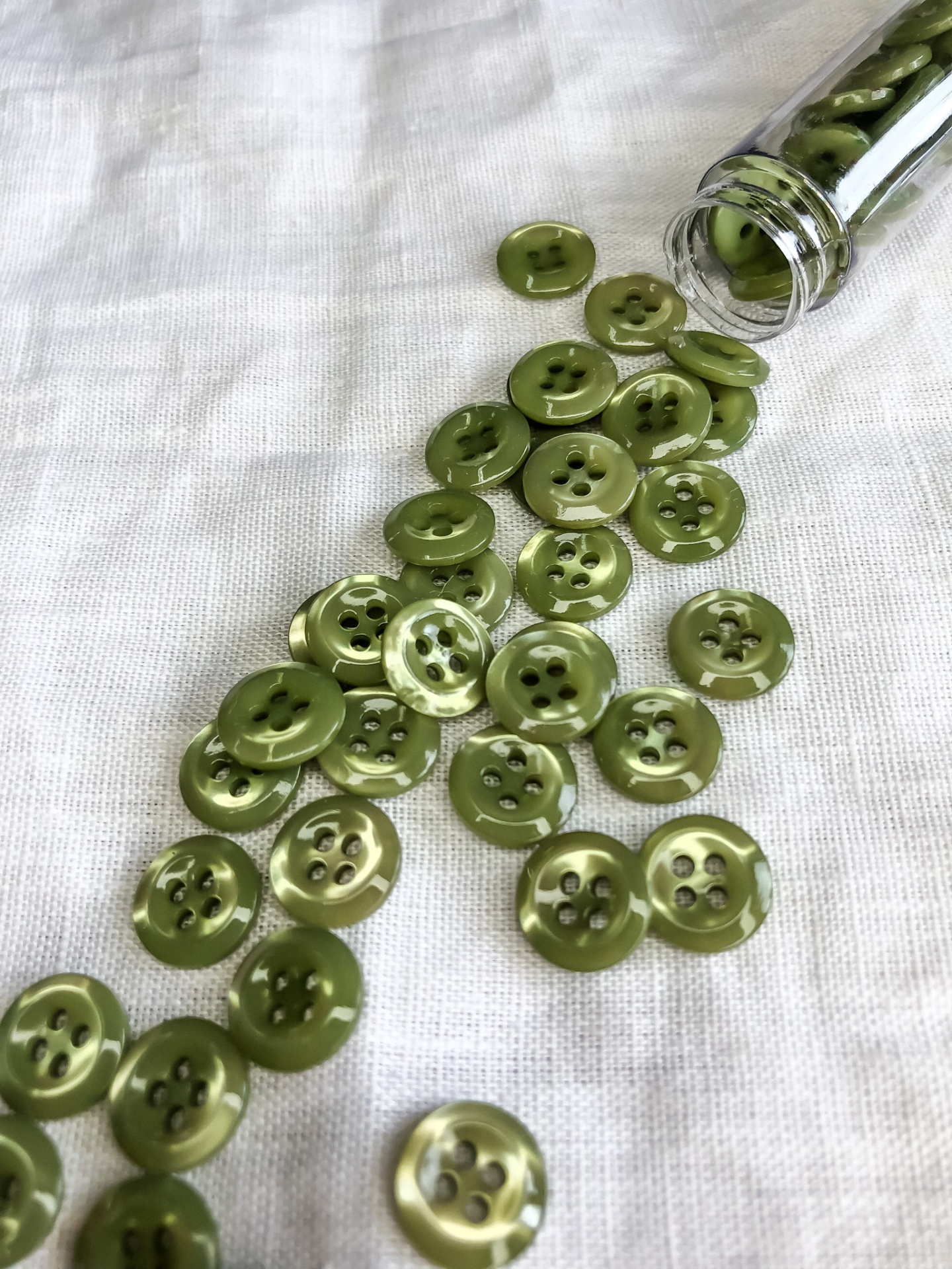 Botões verdes redondos com quatro furos espalhados ao lado de um frasco de vidro sobre um tecido branco