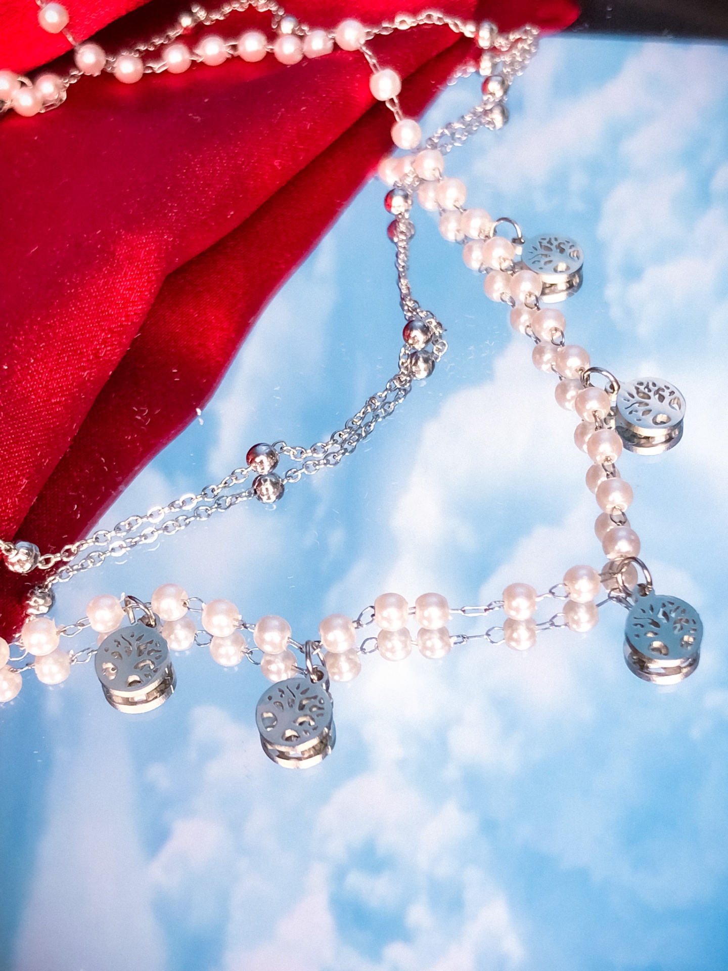 Colar prateado com contas tipo pérola e pendentes redondos sobre espelho refletindo céu azul